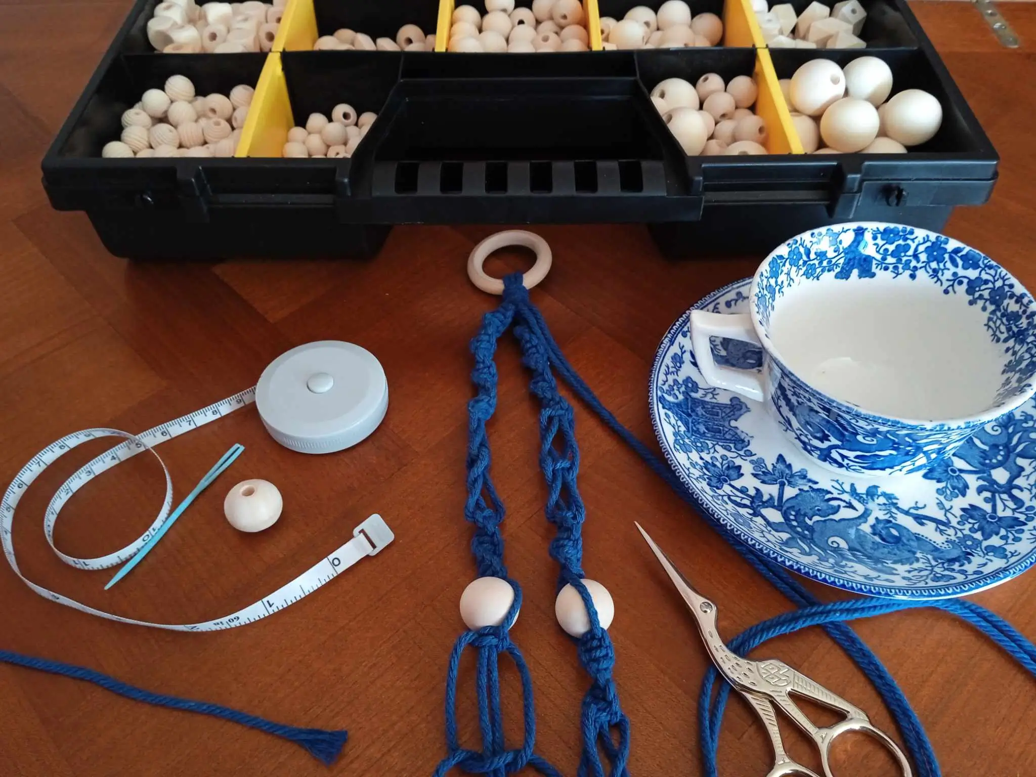 Une caisse contenant des perles en bois, un mètre ruban, une tasses, une paire de ciseaux et une structure en macramé, tout pour le fait main
